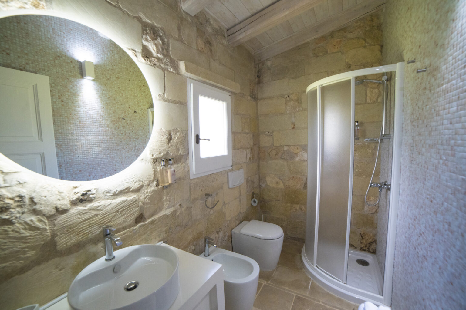 Modern bathroom interior featuring a round illuminated mirror, stone walls, a glass shower enclosure, a white sink, and a toilet. Natural light enters through a window, enhancing the spacious and stylish design. | Only on SopranoVillas