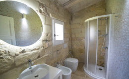Modern bathroom interior featuring a round illuminated mirror, stone walls, a glass shower enclosure, a white sink, and a toilet. Natural light enters through a window, enhancing the spacious and stylish design.