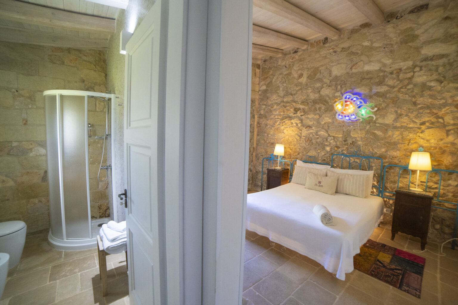 Cozy bedroom with stone walls featuring a double bed, bedside tables, and decorative lighting, alongside a modern bathroom with a glass shower and toilet, creating a serene and stylish retreat. | Only on SopranoVillas