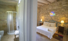 Cozy bedroom with stone walls featuring a double bed, bedside tables, and decorative lighting, alongside a modern bathroom with a glass shower and toilet, creating a serene and stylish retreat.