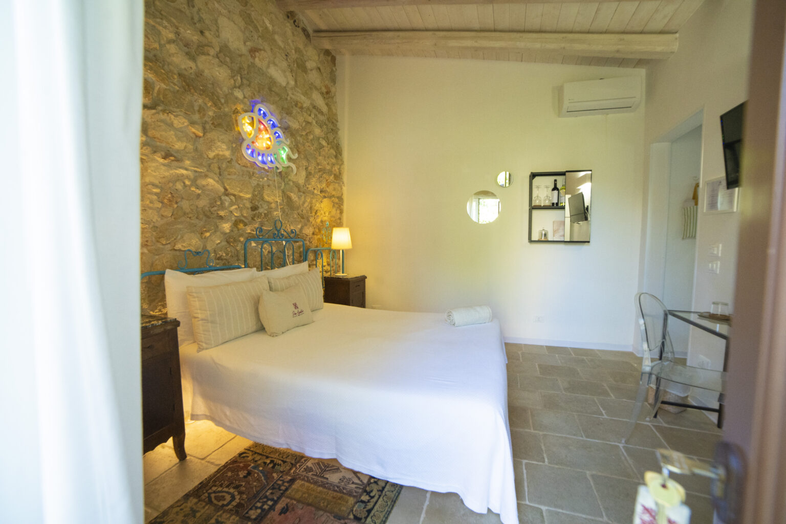 Cozy bedroom featuring a stone wall, a blue metal bed frame, and soft white bedding. The room includes a stylish lamp, a small table, and decorative mirrors, creating a warm and inviting atmosphere. Ideal for a relaxing getaway. | Only on SopranoVillas