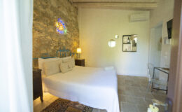 Cozy bedroom featuring a stone wall, a blue metal bed frame, and soft white bedding. The room includes a stylish lamp, a small table, and decorative mirrors, creating a warm and inviting atmosphere. Ideal for a relaxing getaway.