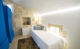 Cozy bedroom featuring a single bed, stone wall accents, modern lighting, and a decorative rug, ideal for a minimalist and stylish retreat.