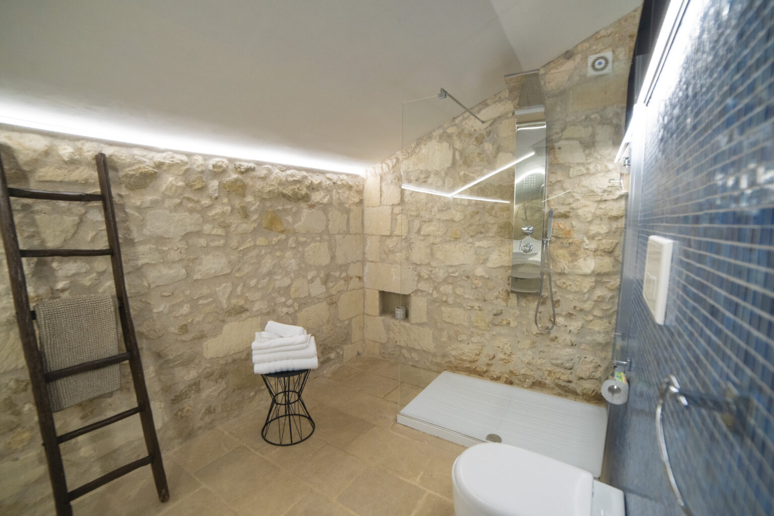 Modern bathroom featuring natural stone walls, a sleek blue tiled shower area, a minimalist white shower base, a large mirror, and neatly stacked white towels on a stylish black stool. The space is illuminated with soft LED lighting, creating a warm and inviting atmosphere. | Only on SopranoVillas