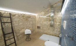 Modern bathroom featuring natural stone walls, a sleek blue tiled shower area, a minimalist white shower base, a large mirror, and neatly stacked white towels on a stylish black stool. The space is illuminated with soft LED lighting, creating a warm and inviting atmosphere.