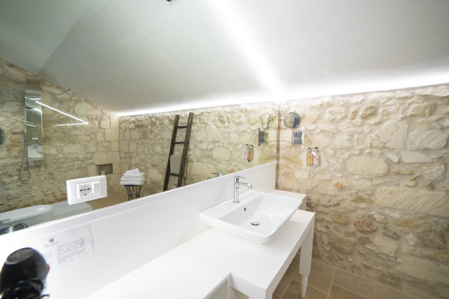Modern bathroom interior featuring a sleek white sink, a large mirror, and stone walls, illuminated by LED lighting. A wooden ladder leans against the wall, adding a rustic touch to the contemporary design. | Only on SopranoVillas