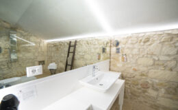 Modern bathroom interior featuring a sleek white sink, a large mirror, and stone walls, illuminated by LED lighting. A wooden ladder leans against the wall, adding a rustic touch to the contemporary design.