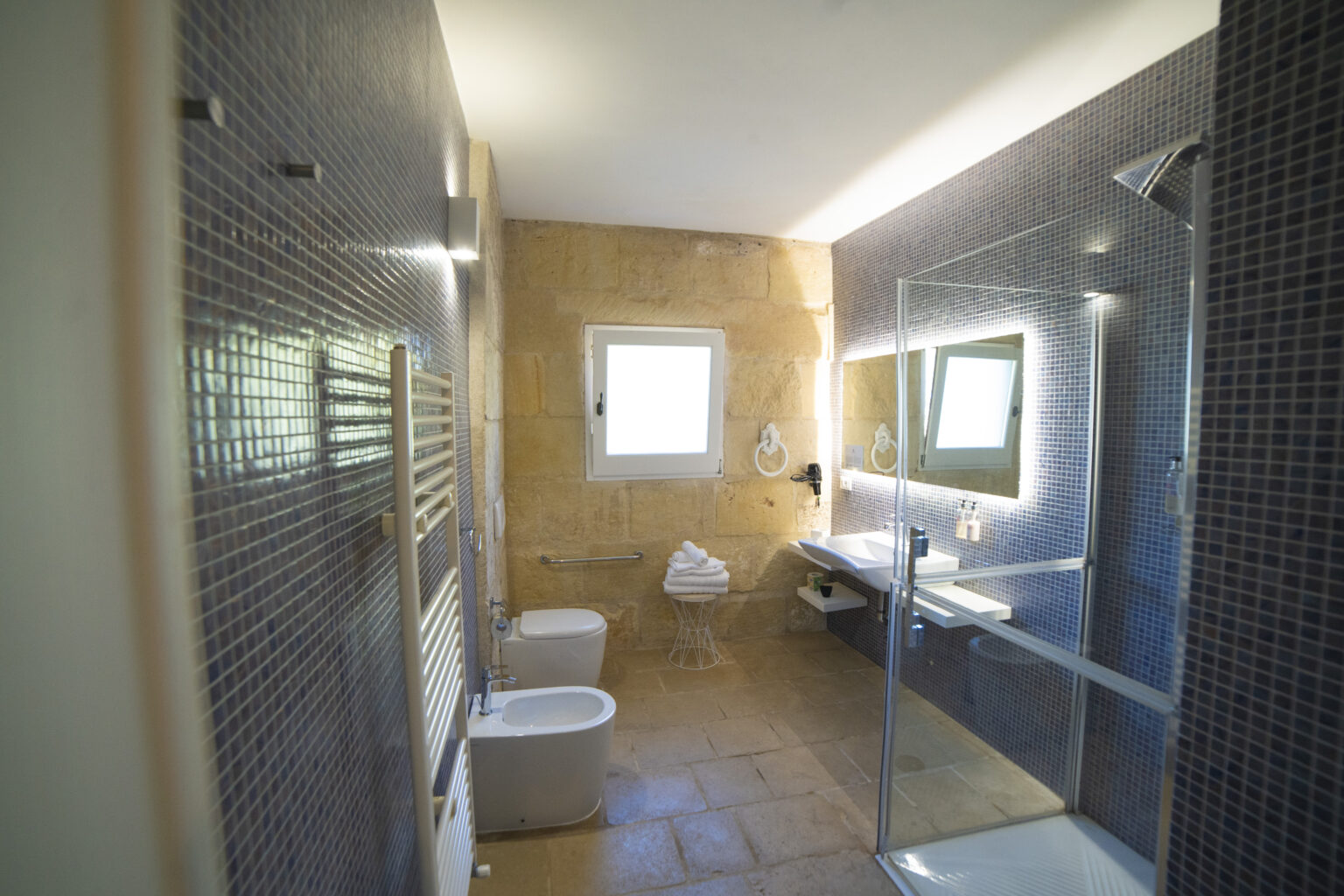 Modern bathroom interior featuring a glass shower, contemporary fixtures, and natural light from a window. The space includes a toilet, bidet, and a sleek vanity with a large mirror, accented by textured wall tiles and warm lighting. | Only on SopranoVillas