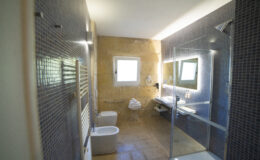 Modern bathroom interior featuring a glass shower, contemporary fixtures, and natural light from a window. The space includes a toilet, bidet, and a sleek vanity with a large mirror, accented by textured wall tiles and warm lighting.