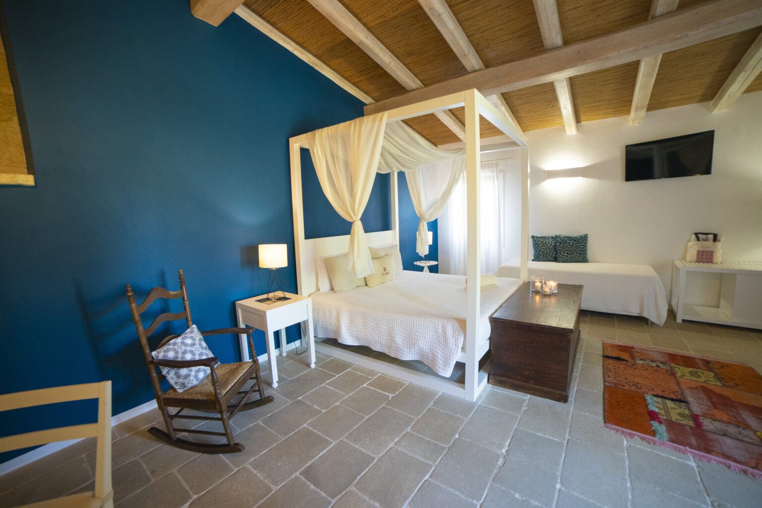 Cozy bedroom featuring a four-poster bed with white drapes, a wooden rocking chair, and a decorative rug. The room is adorned with a blue accent wall and has natural light from a window, creating a serene and inviting atmosphere. | Only on SopranoVillas