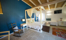 Cozy bedroom featuring a four-poster bed with white drapes, a wooden rocking chair, and a decorative rug. The room is adorned with a blue accent wall and has natural light from a window, creating a serene and inviting atmosphere.