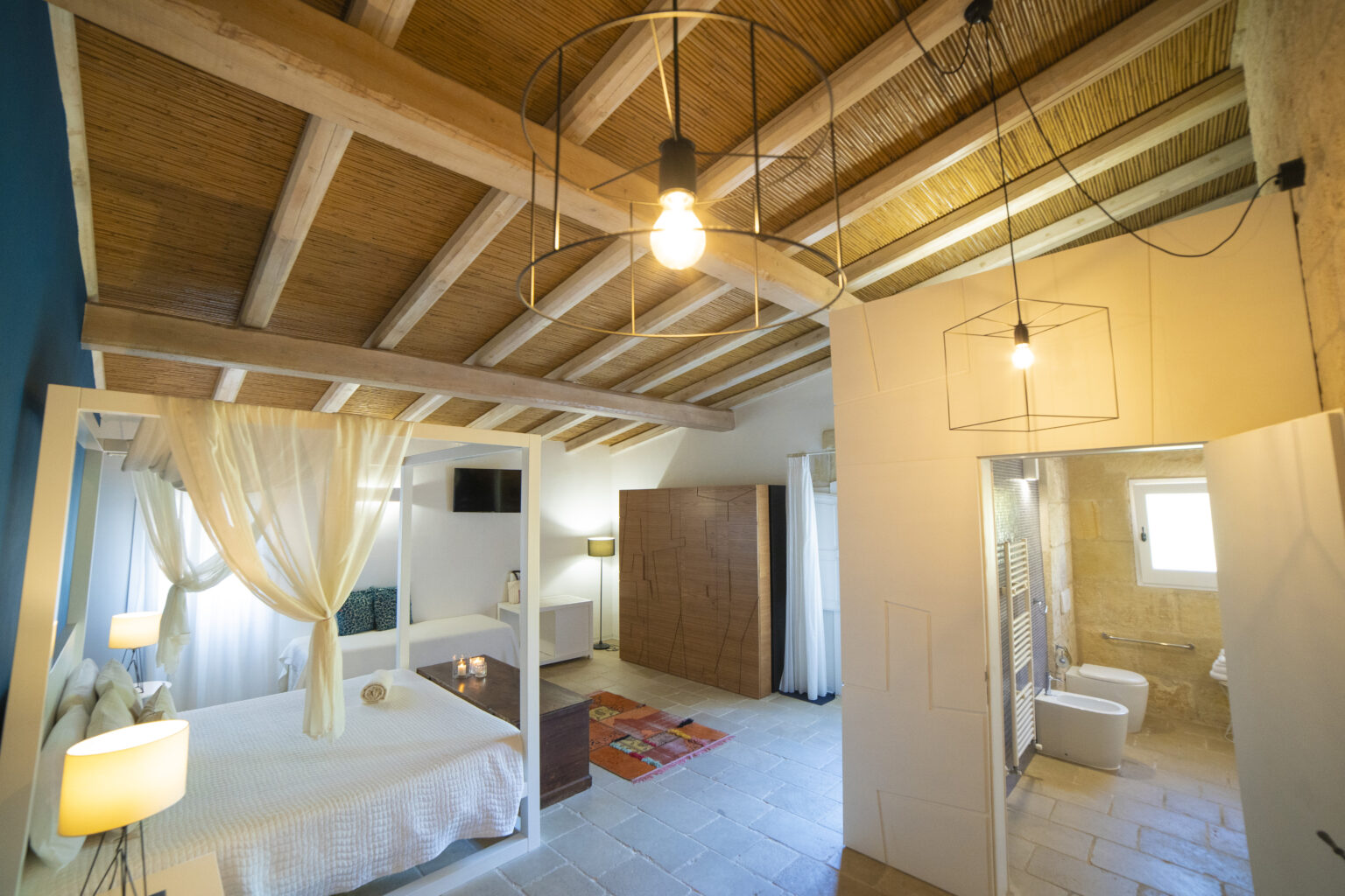 Modern bedroom interior with a four-poster bed, wooden ceiling beams, contemporary lighting, and an adjoining bathroom. The space features neutral tones, elegant decor, and large windows allowing natural light. Ideal for relaxation and comfort in a stylish setting. | Only on SopranoVillas