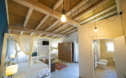 Modern bedroom interior with a four-poster bed, wooden ceiling beams, contemporary lighting, and an adjoining bathroom. The space features neutral tones, elegant decor, and large windows allowing natural light. Ideal for relaxation and comfort in a stylish setting.