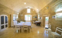 Spacious dining area with high arched stone ceilings, featuring a large table surrounded by white chairs, natural light streaming through windows, and tasteful decor.