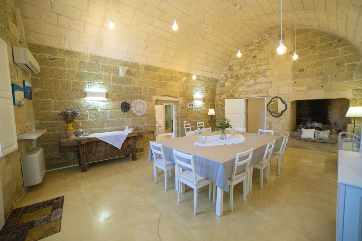 Spacious dining area with a large table surrounded by white chairs, featuring a rustic wooden side table and stone walls, illuminated by pendant lights, creating a warm and inviting atmosphere. | Only on SopranoVillas