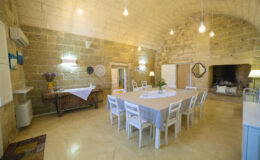 Spacious dining area with a large table surrounded by white chairs, featuring a rustic wooden side table and stone walls, illuminated by pendant lights, creating a warm and inviting atmosphere.