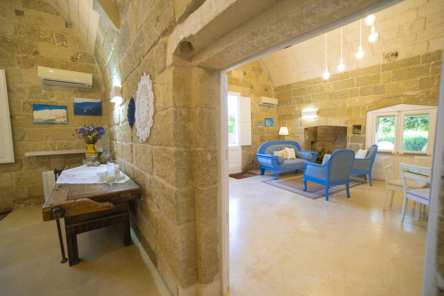 Interior view of a cozy living space featuring stone walls, a wooden dining table, and blue furniture. The room is illuminated by hanging lights and includes decorative artwork on the walls, creating a warm and inviting atmosphere. | Only on SopranoVillas