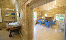 Interior view of a cozy living space featuring stone walls, a wooden dining table, and blue furniture. The room is illuminated by hanging lights and includes decorative artwork on the walls, creating a warm and inviting atmosphere.