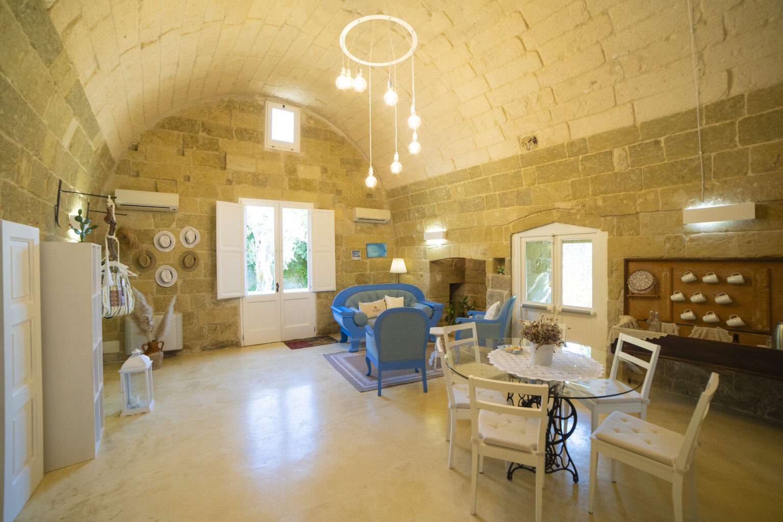 Cozy living space with a stone arched ceiling, featuring blue furniture, a glass dining table, and decorative elements like hats and plants. Natural light streams in through large windows, enhancing the inviting atmosphere. Perfect for relaxation or entertaining guests. | Only on SopranoVillas