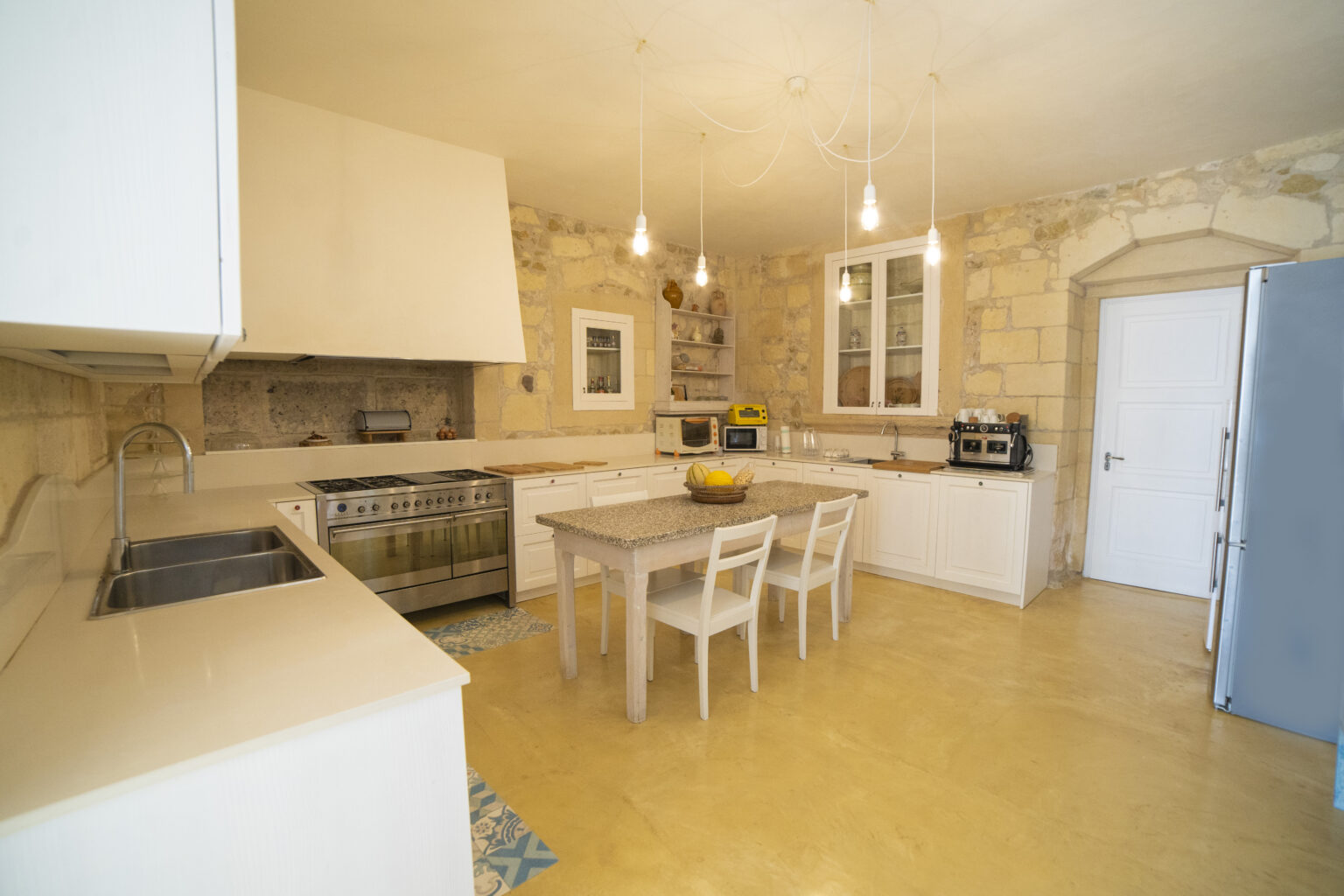 Bright and modern kitchen featuring white cabinetry, a large island with a granite countertop, stainless steel appliances, and natural stone walls. The space includes a dining table with white chairs and various kitchen gadgets, illuminated by stylish pendant lights. | Only on SopranoVillas