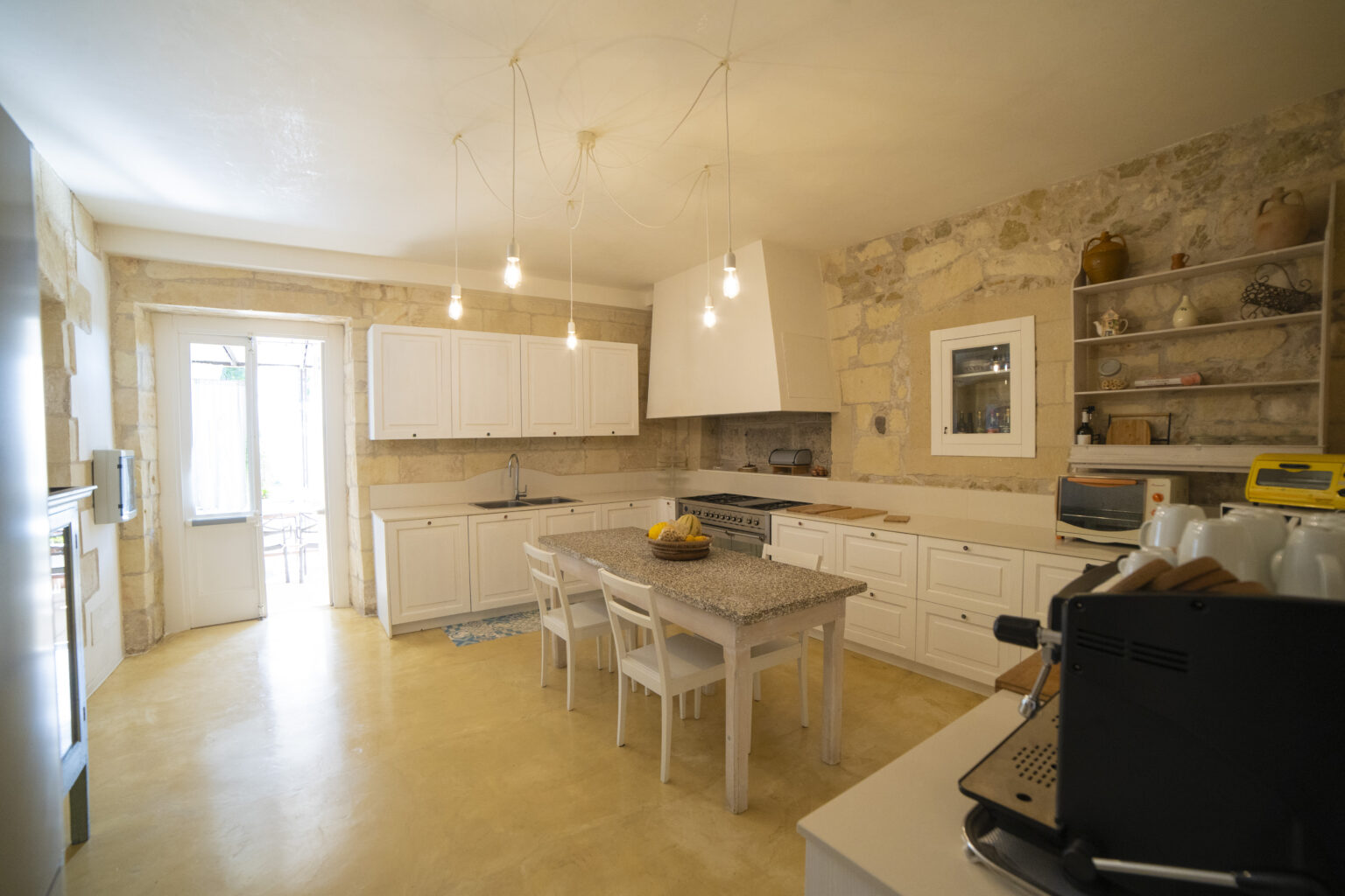 Modern kitchen interior featuring white cabinetry, a granite island, and stone walls. The space is illuminated by hanging light bulbs and includes a stainless steel stove and a cozy dining area with white chairs. Natural light streams in through the open door, enhancing the inviting atmosphere. | Only on SopranoVillas