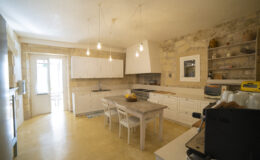 Modern kitchen interior featuring white cabinetry, a granite island, and stone walls. The space is illuminated by hanging light bulbs and includes a stainless steel stove and a cozy dining area with white chairs. Natural light streams in through the open door, enhancing the inviting atmosphere.