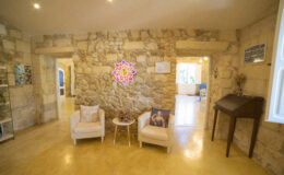 Cozy interior of a stone-walled living space featuring two armchairs, a small table, and decorative wall art. Natural lighting enhances the warm ambiance, creating an inviting atmosphere.