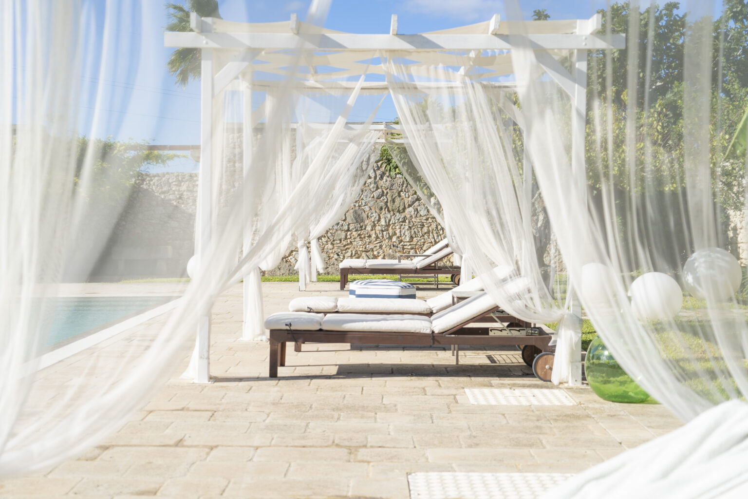 A serene outdoor lounging area featuring a wooden sunbed draped with sheer white curtains, overlooking a swimming pool surrounded by lush greenery. Perfect for relaxation and enjoying sunny days. | Only on SopranoVillas