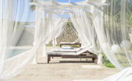 A serene outdoor lounging area featuring a wooden sunbed draped with sheer white curtains, overlooking a swimming pool surrounded by lush greenery. Perfect for relaxation and enjoying sunny days.