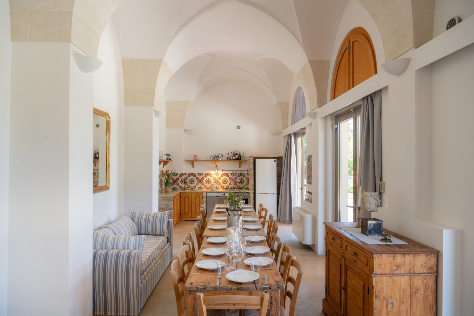 Bright and airy dining area featuring a long wooden table set for a meal, surrounded by rustic wooden chairs. The room showcases high arched ceilings, a cozy striped sofa, and a decorative tiled wall. Natural light floods in through large windows, enhancing the inviting atmosphere. | Only on SopranoVillas
