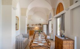 Bright and airy dining area featuring a long wooden table set for a meal, surrounded by rustic wooden chairs. The room showcases high arched ceilings, a cozy striped sofa, and a decorative tiled wall. Natural light floods in through large windows, enhancing the inviting atmosphere.