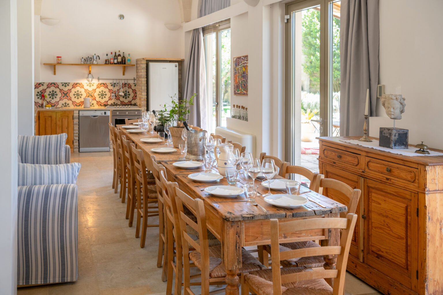 Bright and spacious dining area featuring a long wooden table set for a meal, surrounded by wooden chairs. The open kitchen showcases modern appliances and colorful tiled backsplash, while large windows provide natural light and views of the outdoor garden. Cozy seating area with striped chairs enhances the inviting atmosphere. | Only on SopranoVillas