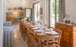 Bright and spacious dining area featuring a long wooden table set for a meal, surrounded by wooden chairs. The open kitchen showcases modern appliances and colorful tiled backsplash, while large windows provide natural light and views of the outdoor garden. Cozy seating area with striped chairs enhances the inviting atmosphere.