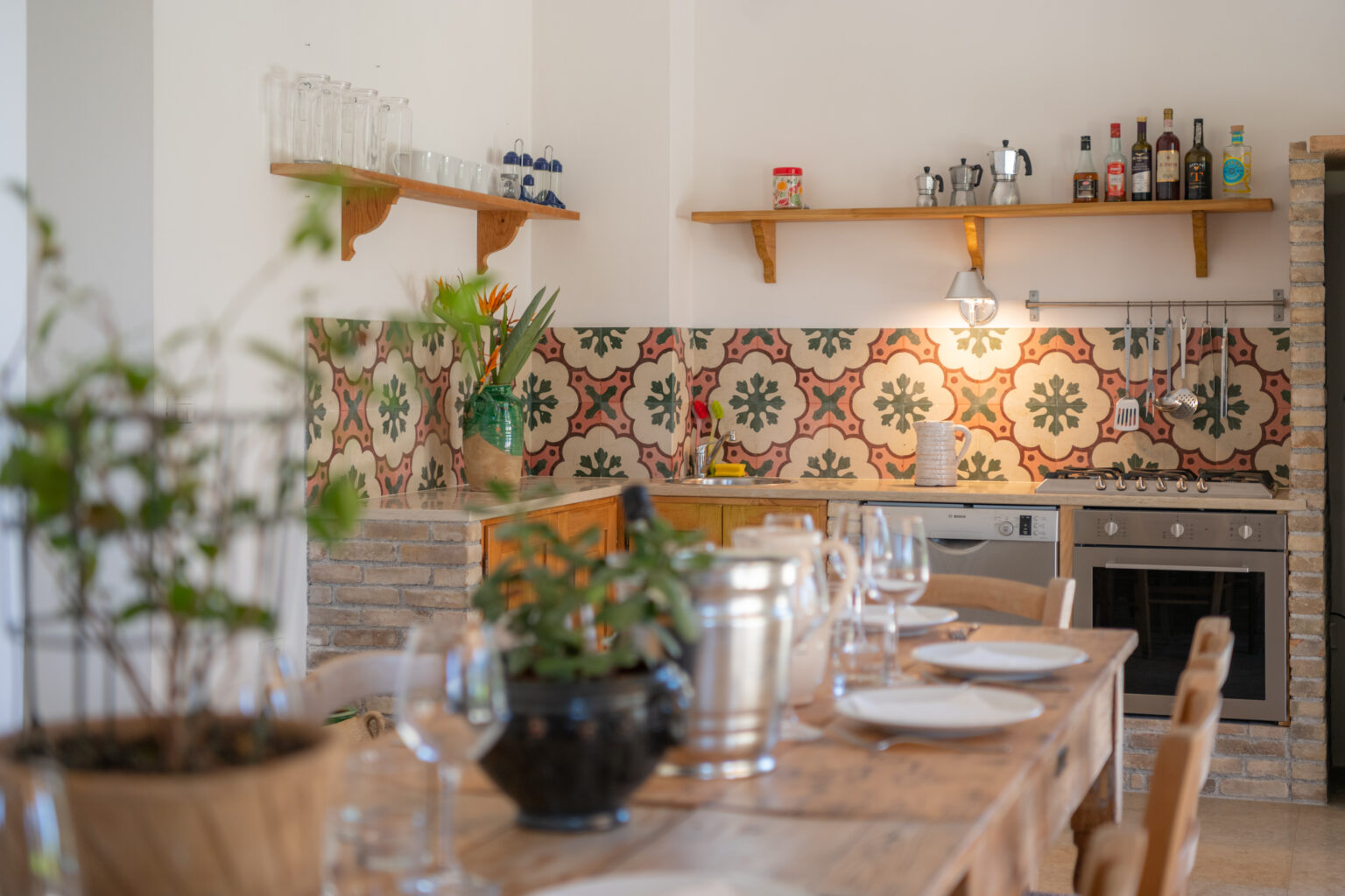 Bright and inviting kitchen interior featuring wooden cabinetry, patterned tile backsplash, and a rustic dining table set with plates and glassware. The space includes decorative plants, kitchen appliances, and a shelf displaying glassware and bottles, creating a warm and welcoming atmosphere. | Only on SopranoVillas