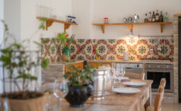 Bright and inviting kitchen interior featuring wooden cabinetry, patterned tile backsplash, and a rustic dining table set with plates and glassware. The space includes decorative plants, kitchen appliances, and a shelf displaying glassware and bottles, creating a warm and welcoming atmosphere.
