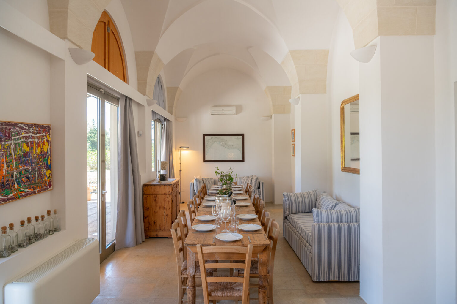 Dining area with a long wooden table set for a meal, featuring rustic chairs, a striped sofa, and large windows allowing natural light. Decor includes a colorful painting and a vintage map on the wall, complemented by soft curtains and a wooden sideboard. | Only on SopranoVillas