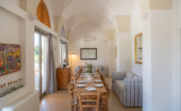 Dining area with a long wooden table set for a meal, featuring rustic chairs, a striped sofa, and large windows allowing natural light. Decor includes a colorful painting and a vintage map on the wall, complemented by soft curtains and a wooden sideboard.