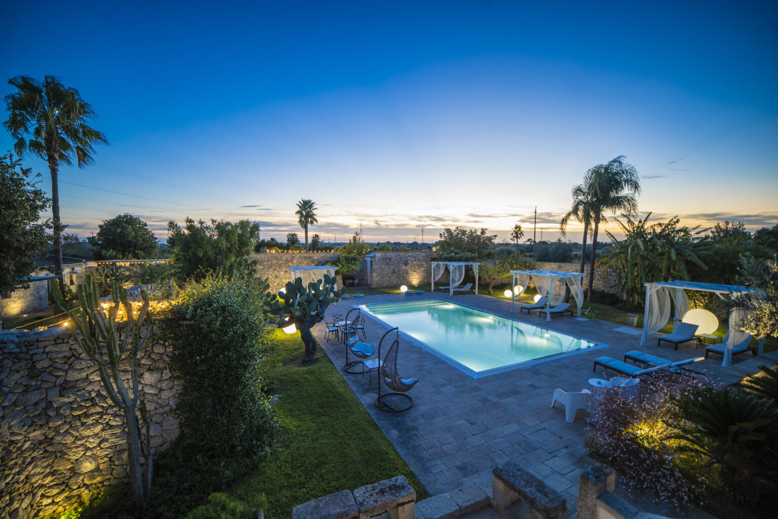 Luxurious outdoor pool area at sunset, featuring palm trees, landscaped gardens, and illuminated seating, perfect for relaxation and entertainment. | Only on SopranoVillas