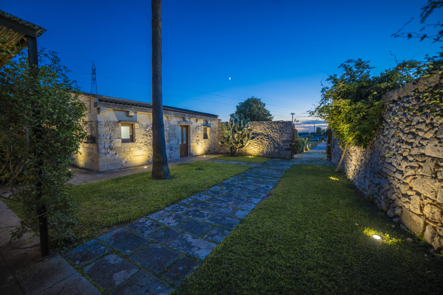 Charming stone pathway leading through a beautifully landscaped garden at dusk, with warm outdoor lighting illuminating the rustic buildings and greenery. | Only on SopranoVillas