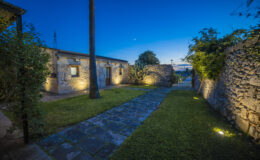 Charming stone pathway leading through a beautifully landscaped garden at dusk, with warm outdoor lighting illuminating the rustic buildings and greenery.