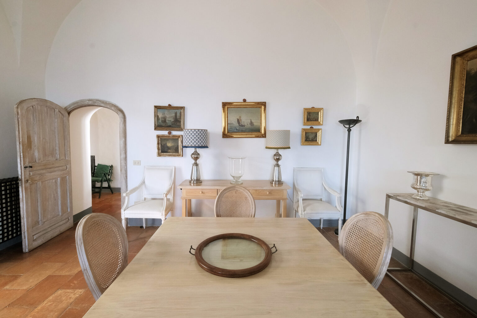 Elegant dining area featuring a wooden table with a decorative tray, surrounded by stylish chairs. The room is adorned with framed artwork on the white walls, illuminated by table lamps and a floor lamp, creating a sophisticated ambiance. | Only on SopranoVillas