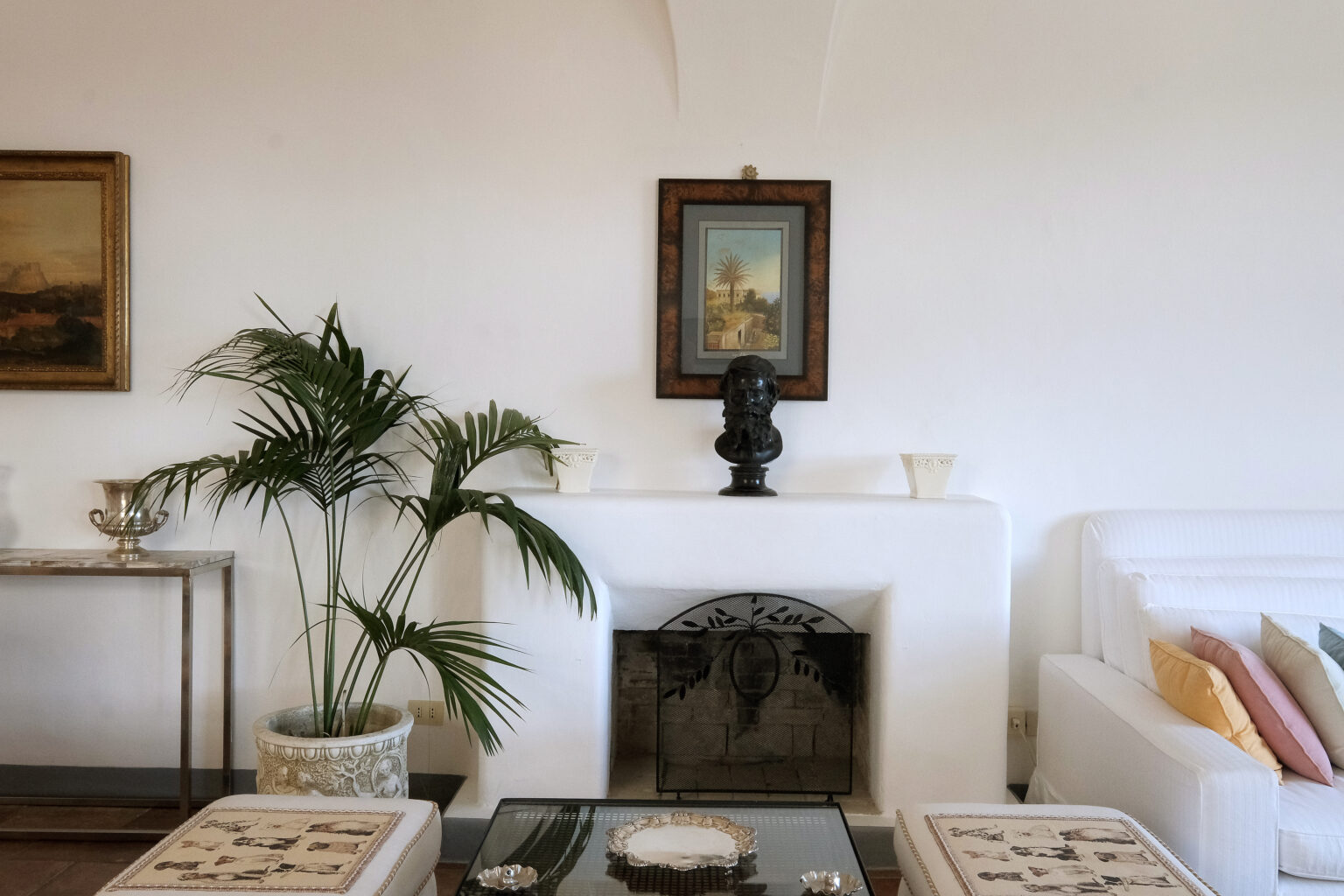 Cozy living room featuring a white sofa with colorful cushions, a decorative plant, and a classic fireplace. Art pieces adorn the walls, including a framed painting above a black bust sculpture. Elegant coffee table with decorative items completes the inviting atmosphere. | Only on SopranoVillas