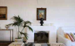 Cozy living room featuring a white sofa with colorful cushions, a decorative plant, and a classic fireplace. Art pieces adorn the walls, including a framed painting above a black bust sculpture. Elegant coffee table with decorative items completes the inviting atmosphere.