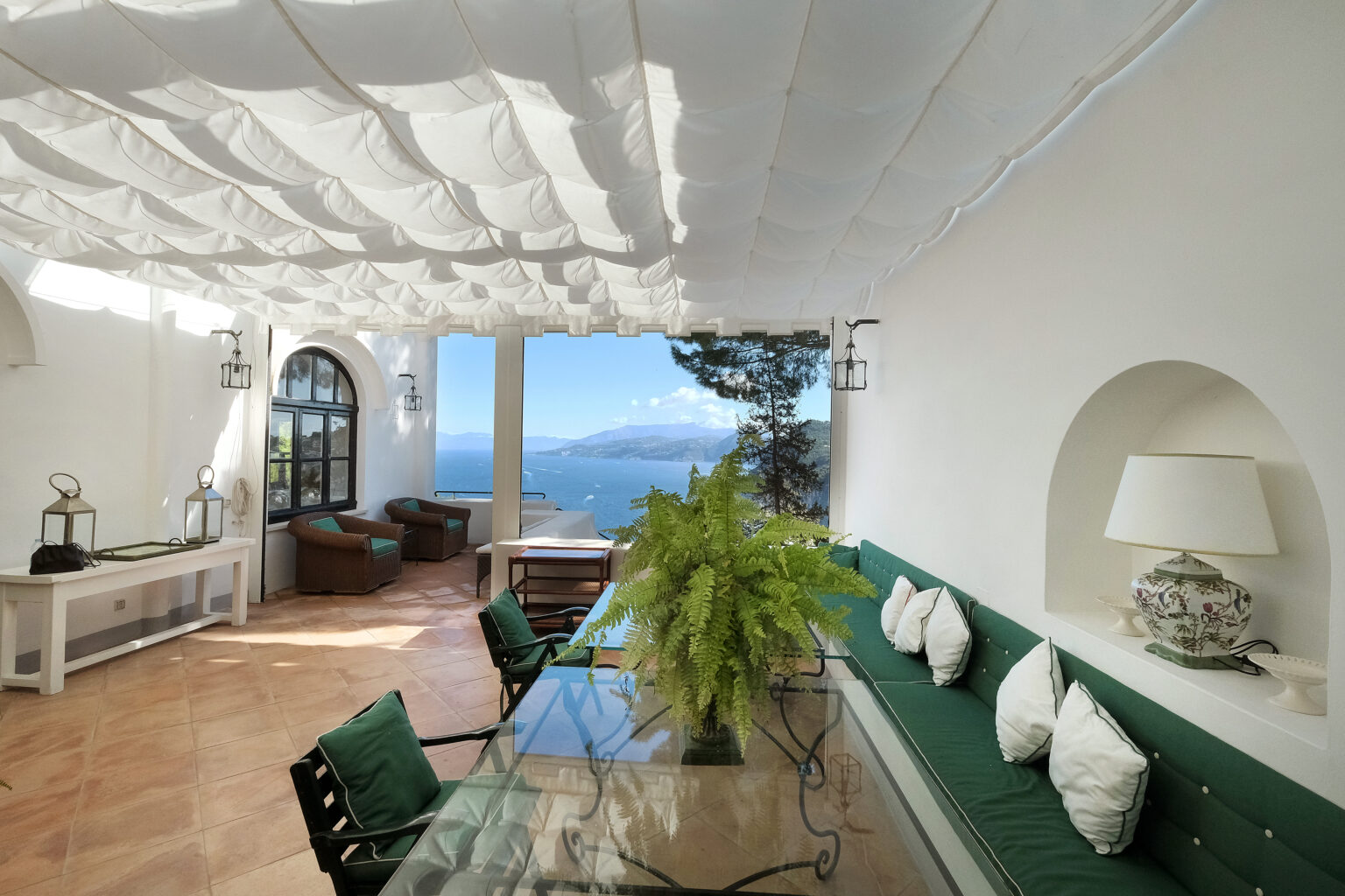 Bright and airy outdoor living space featuring a glass dining table, green upholstered seating, and a stunning ocean view, all under a white draped canopy. Ideal for relaxation and entertaining in a coastal setting. | Only on SopranoVillas