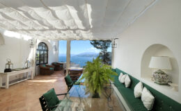 Bright and airy outdoor living space featuring a glass dining table, green upholstered seating, and a stunning ocean view, all under a white draped canopy. Ideal for relaxation and entertaining in a coastal setting.