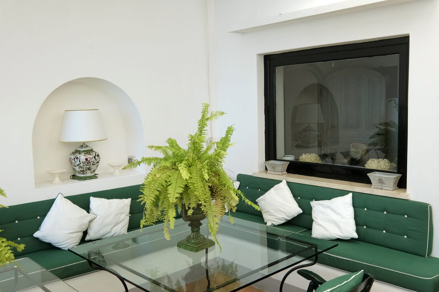 Cozy indoor seating area featuring a green upholstered couch with white pillows, a glass coffee table, and a decorative fern plant. A modern fireplace and wall lamp enhance the inviting atmosphere of the space. | Only on SopranoVillas