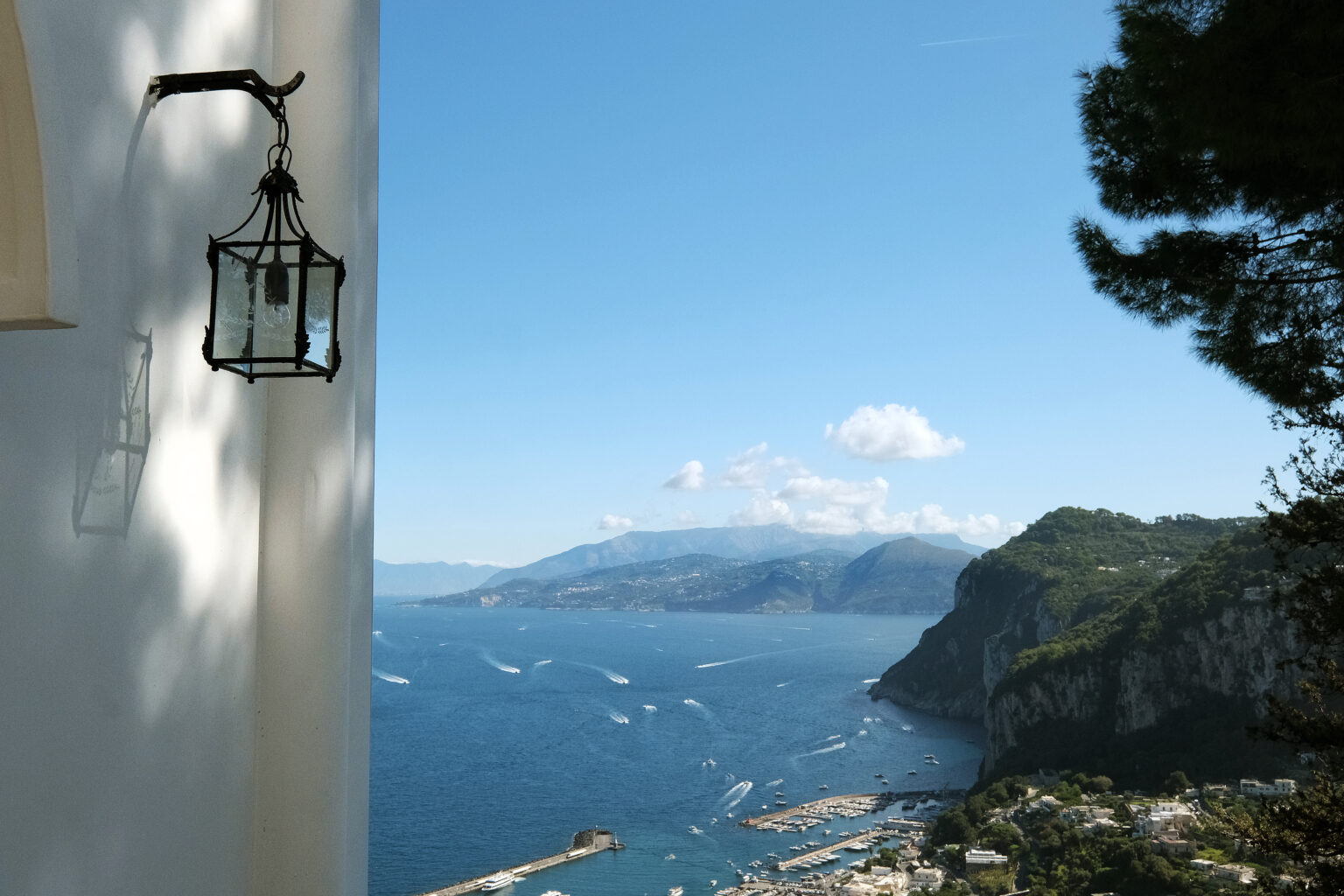 Rustic lantern hanging on a white wall overlooking a serene coastal view with mountains and the sea under a clear blue sky. | Only on SopranoVillas