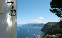 Rustic lantern hanging on a white wall overlooking a serene coastal view with mountains and the sea under a clear blue sky.