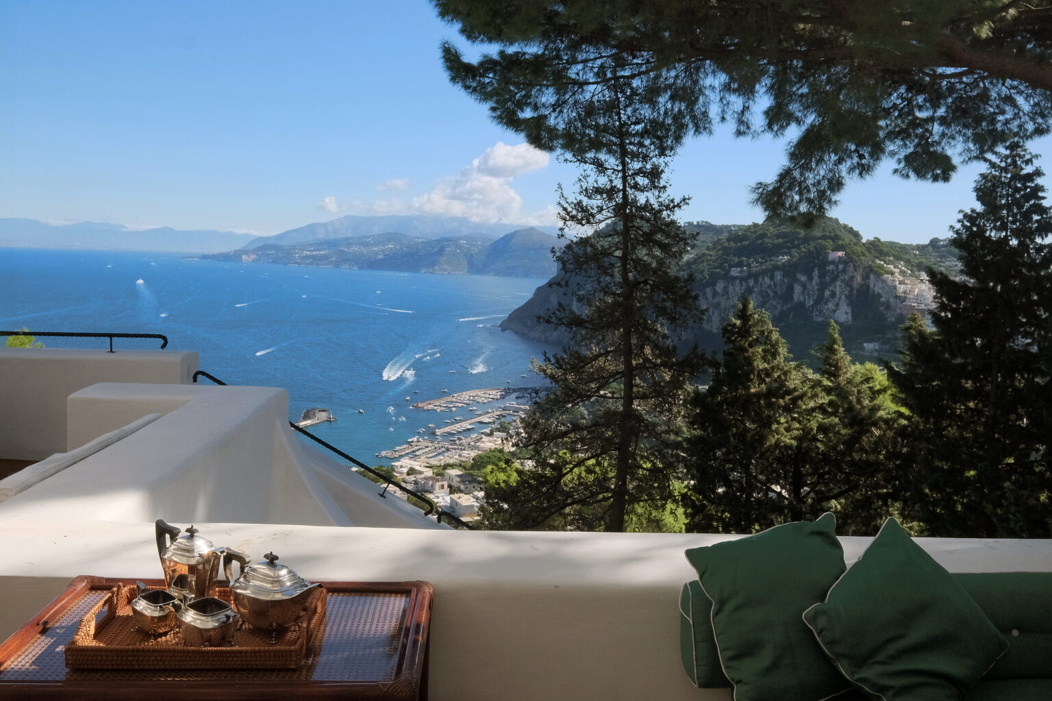 Scenic view of a coastal landscape from a terrace, featuring lush green trees, a tranquil sea, and distant mountains under a clear blue sky, ideal for relaxation and outdoor living. | Only on SopranoVillas