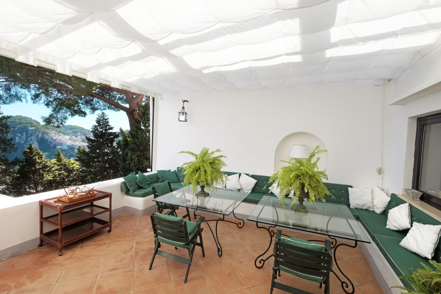Elegant outdoor patio featuring a glass dining table surrounded by green chairs, adorned with potted ferns and cozy cushions. The space is shaded by a white canopy, offering a scenic view of lush trees and mountains in the background, perfect for relaxation and entertaining. | Only on SopranoVillas